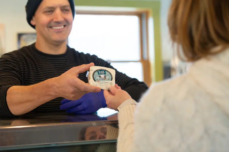 A man stands behind a counter and offers a customer a block of fresh cheese.