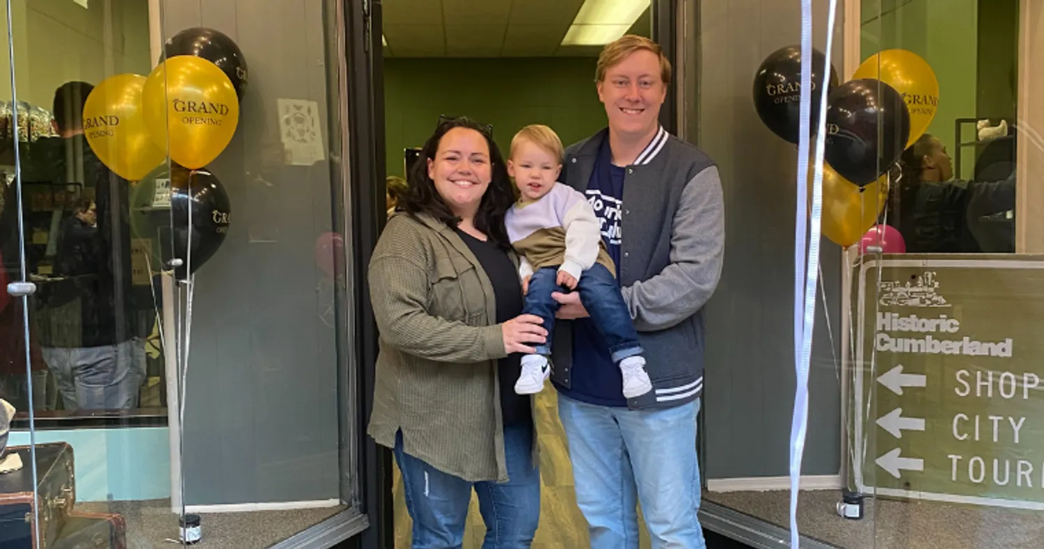 A man, woman, and baby stand outside of a shop, with balloons on either side of them that read "Grand Opening."