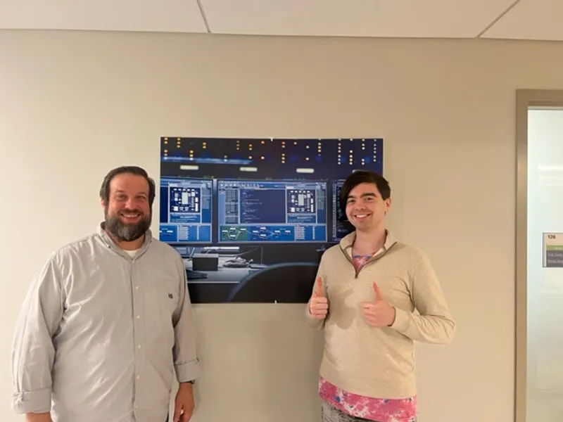 P-Tech intern poses with two-thumbs-up next to their internship supervisor in a white room