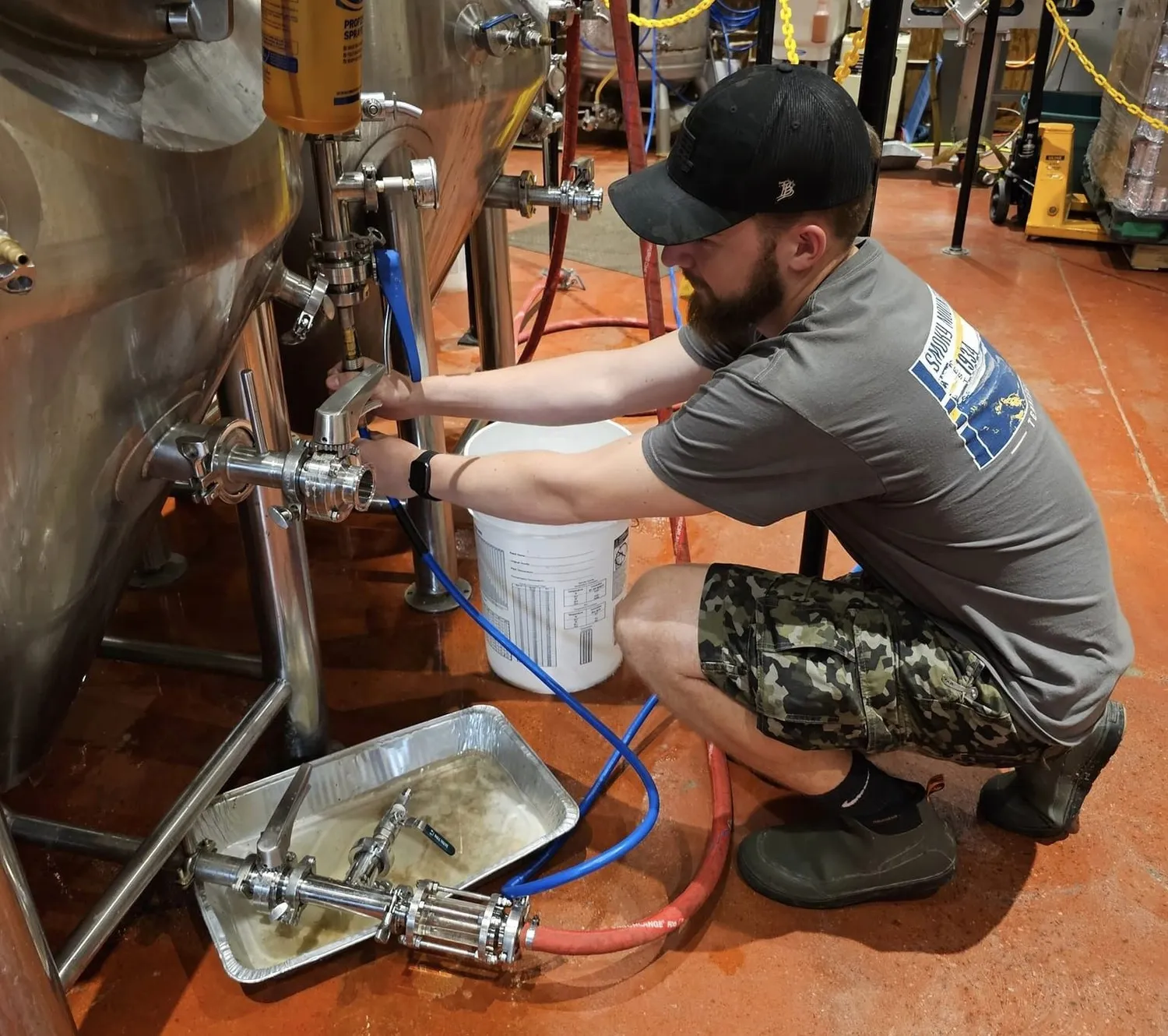 A man in a black hat crouches in front of large beer brewing equipment to pour a sample.