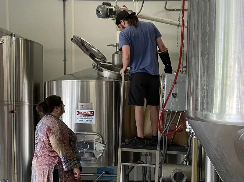 A man and woman work on large silver brewing equipment in a manufacturing setting.