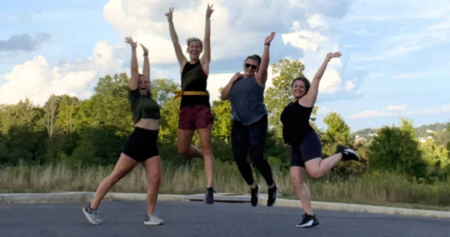 Four women in workout gear jump in the air outdoors.