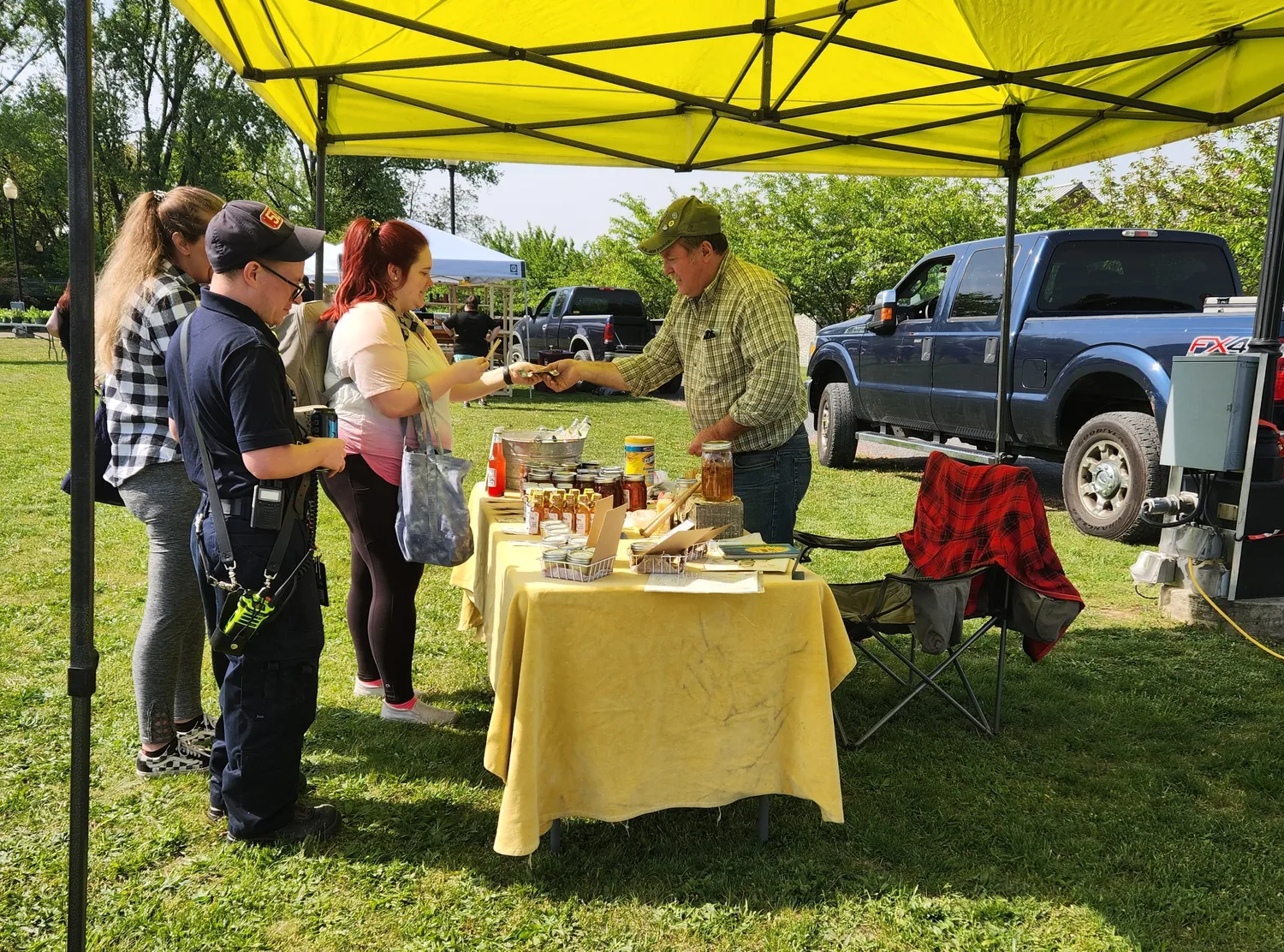 Three people stand at a bright yellow table of a honey seller at the Cumberland Farmers Market.