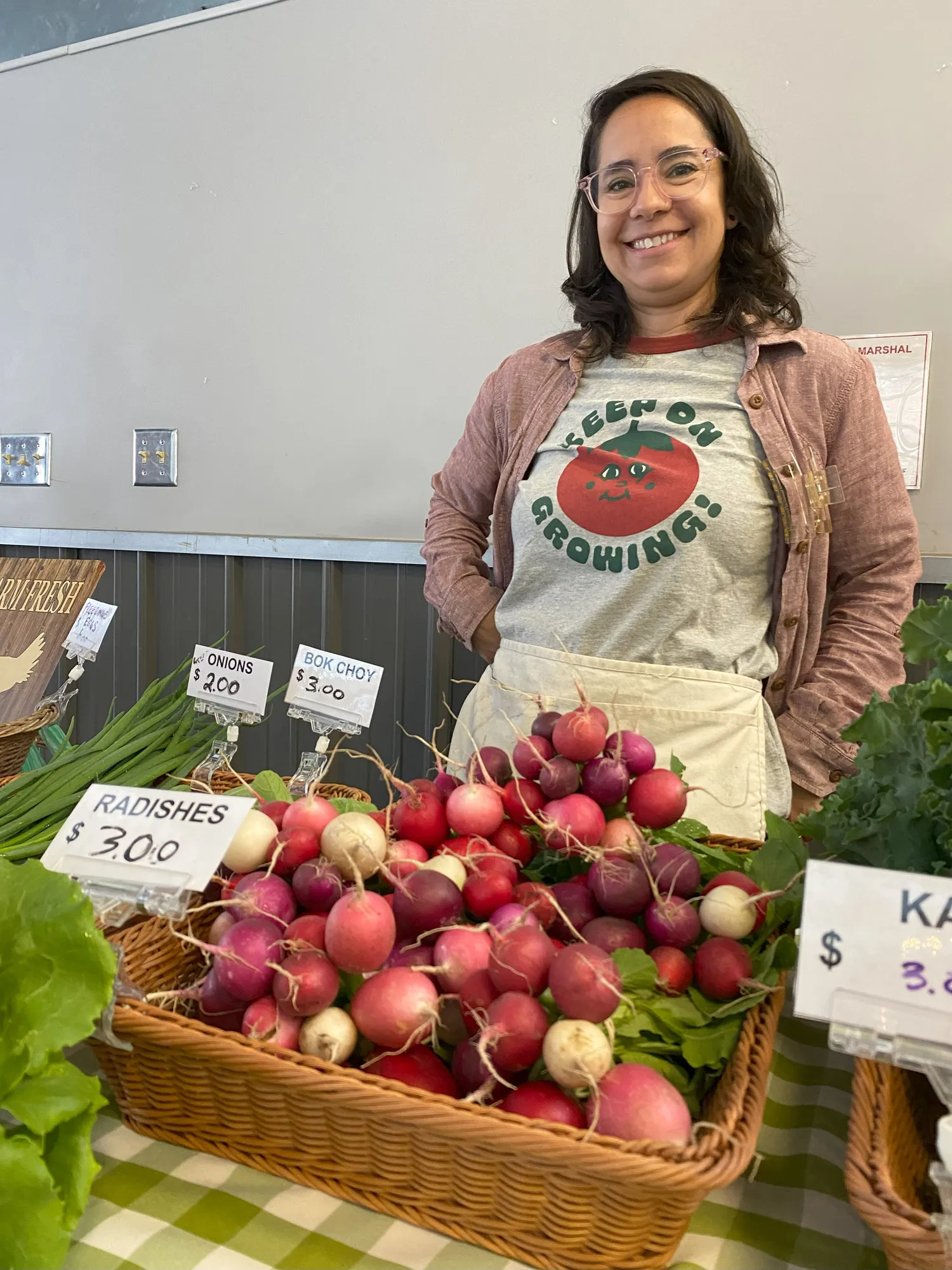 A woman stands behind a table with large selections of beets, onions, and other produce.